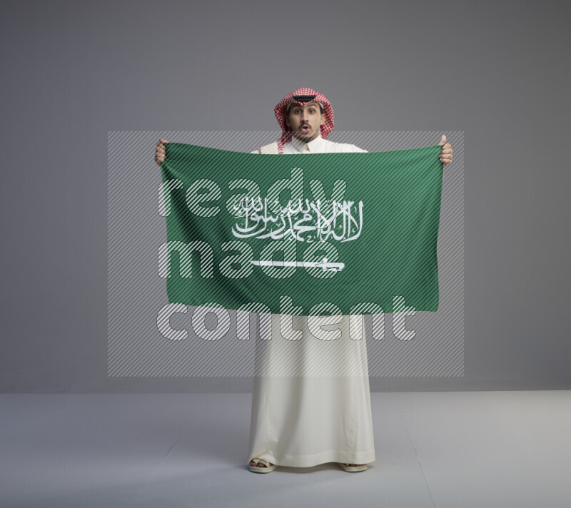 A saudi man standing wearing thob and red shomag holding big saudi flag on gray background