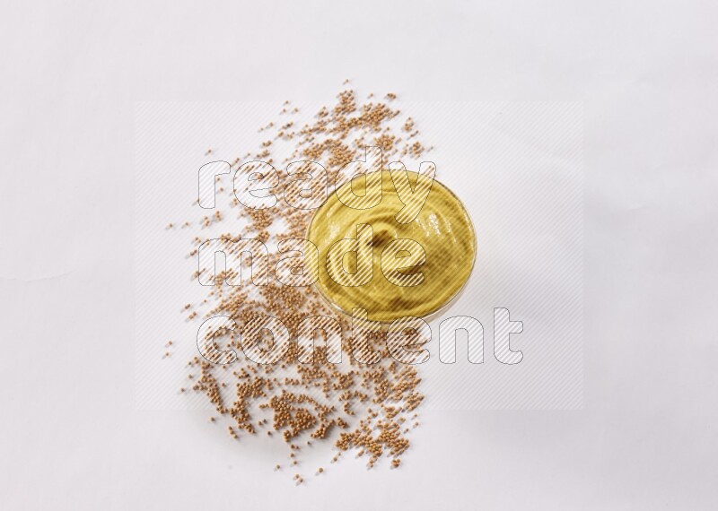 A glass bowl full of mustard paste with mustard seeds underneath on white flooring