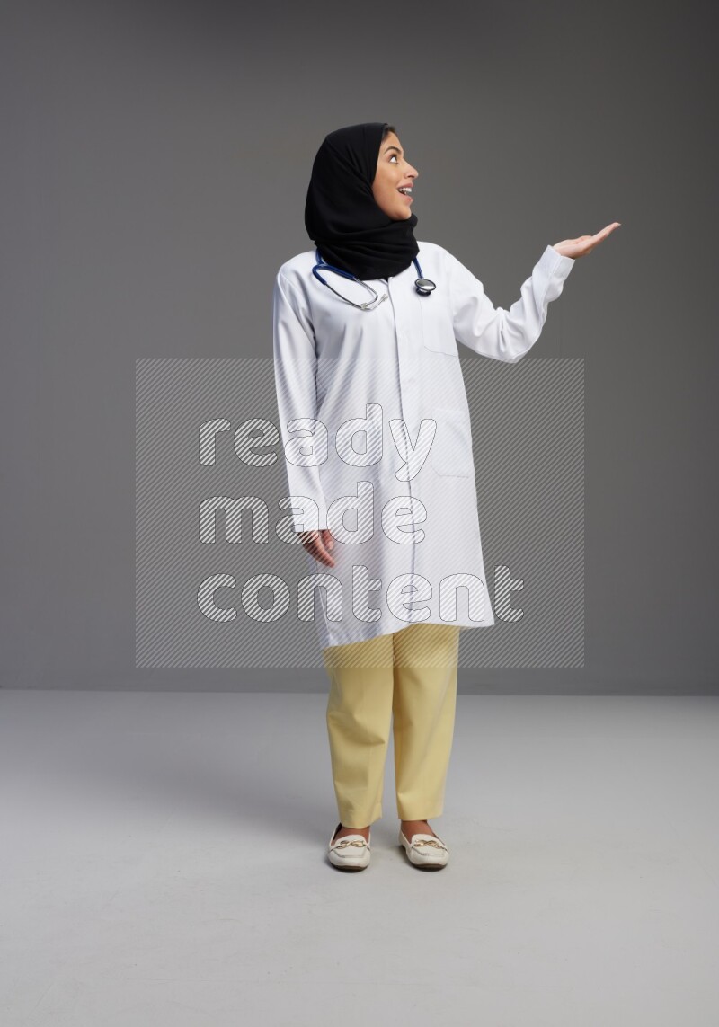 Saudi woman wearing lab coat with stethoscope standing interacting with the camera on Gray background