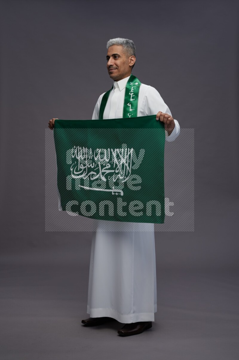 Saudi man wearing thob standing holding Saudi flag on gray background