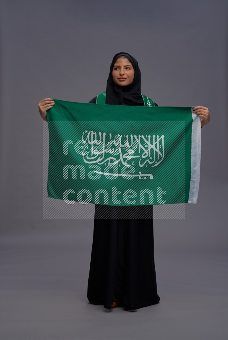Saudi woman wearing Abaya standing holding Saudi flag on gray background