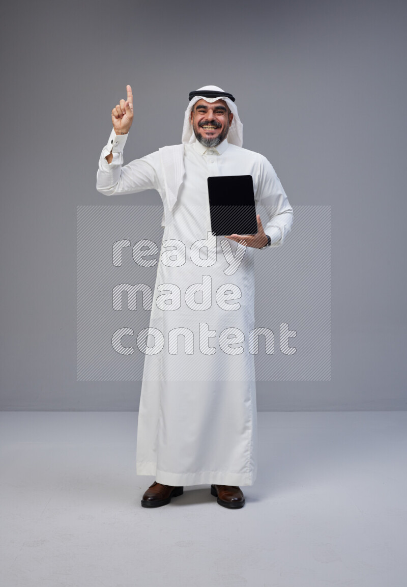 Saudi man Wearing Thob and white Shomag standing showing tablet to camera on Gray background