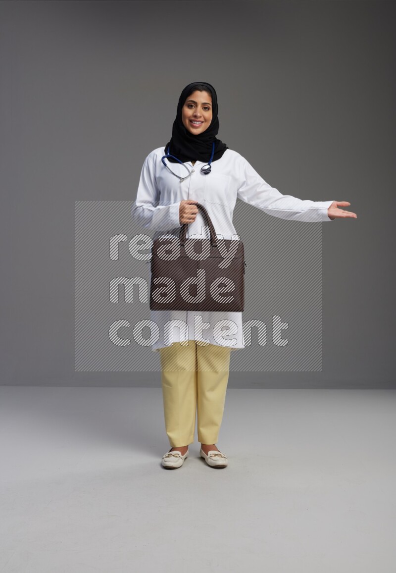Saudi woman wearing lab coat with stethoscope standing holding bag on Gray background