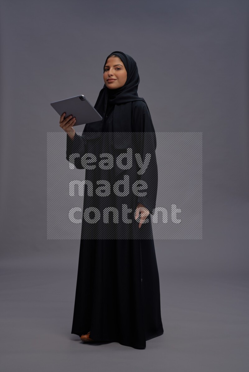Saudi woman wearing Abaya standing working on tablet on gray background