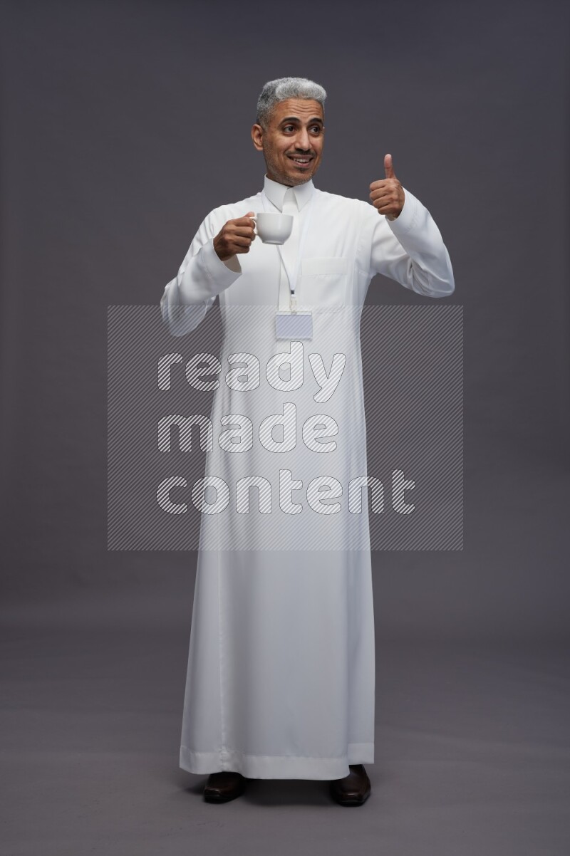 Saudi man wearing thob with neck strap employee badge standing holding mug on gray background