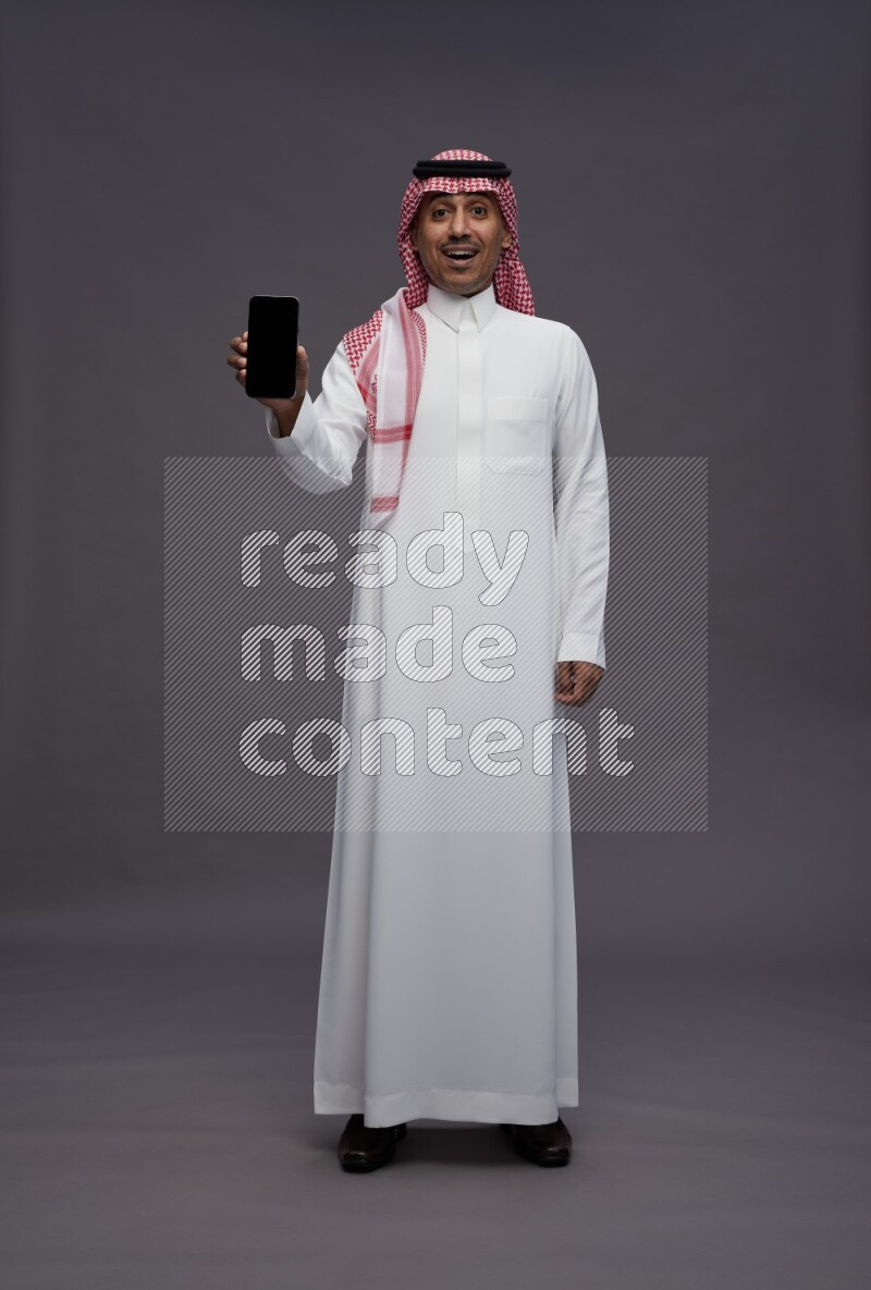 Saudi man wearing thob and shomag standing showing phone to camera on gray background