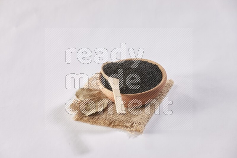 A wooden bowl and wooden spoon full of black seeds on a piece of burlap on a white flooring