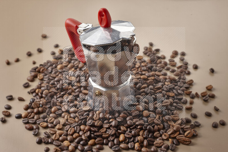 A moka pot with red handle surrounded by roasted coffee beans on beige background