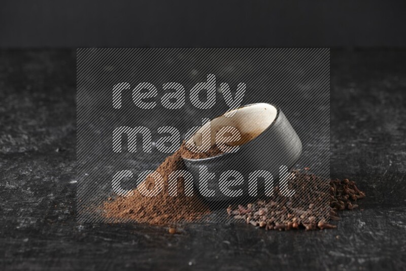 A black pottery bowl full of cloves powder spilled out of it with whole grains on a textured black flooring