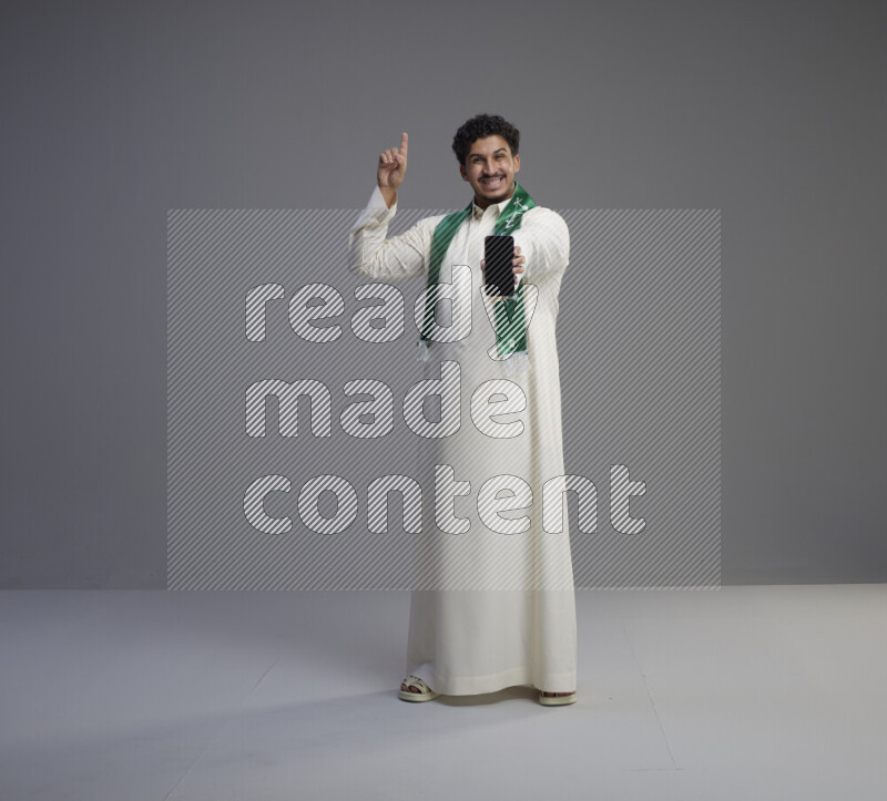 A Saudi man standing wearing thob and Saudi flag scarf showing phone to camera on gray background