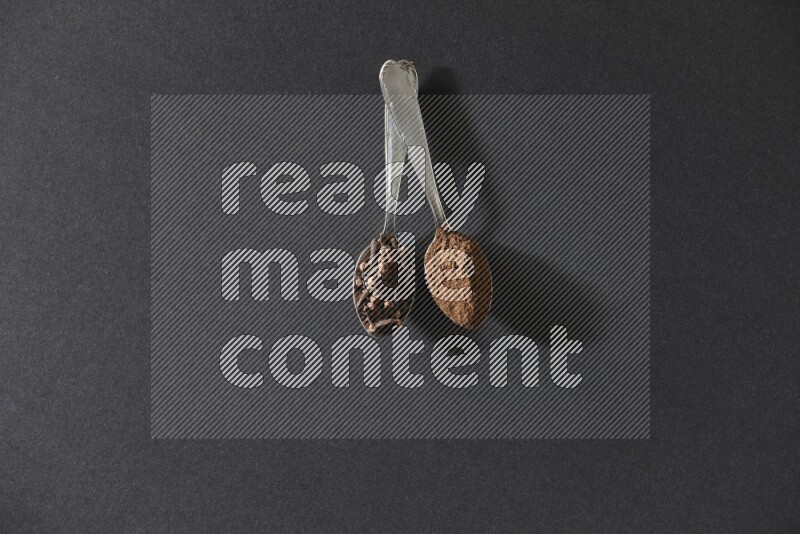 2 metal spoons, one full of cloves grains and the other full of cloves powder on a black flooring