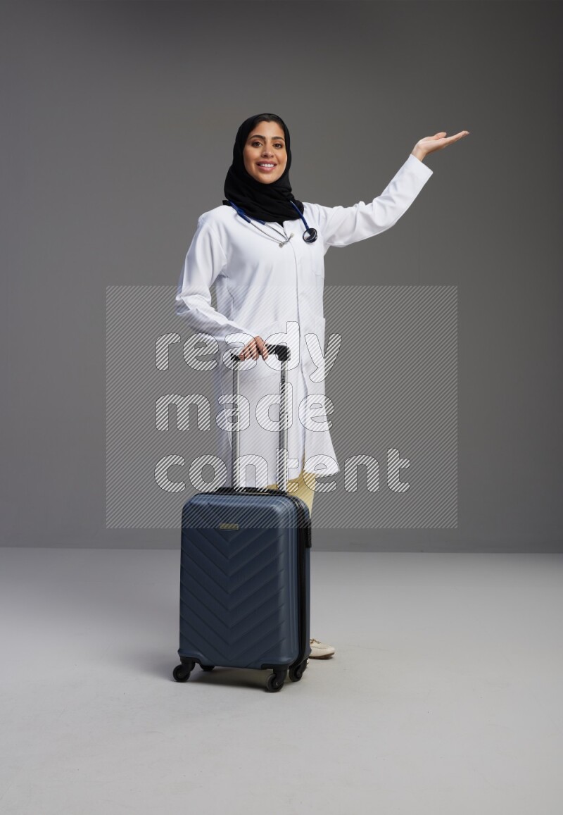 Saudi woman wearing lab coat with stethoscope standing holding Travel bag on Gray background