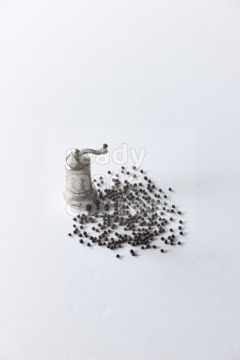 A turkish metal pepper grinder and spreaded black pepper beads on a white flooring