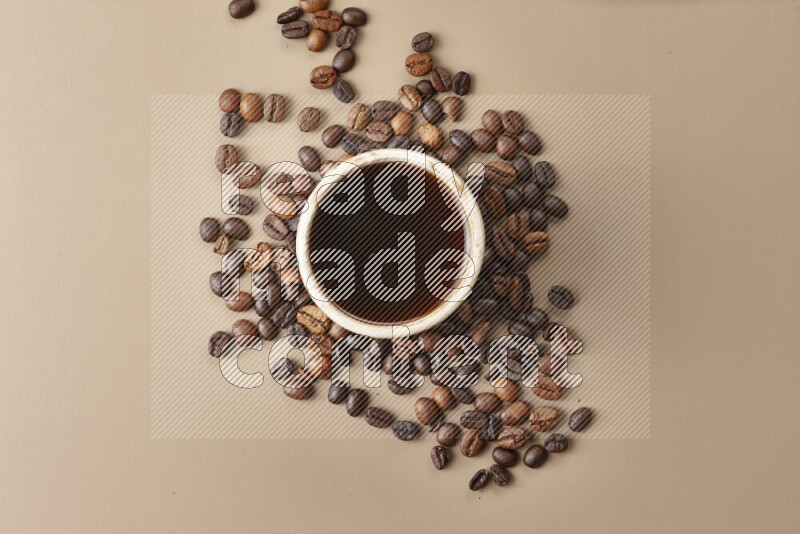 A beige pottery cup of coffee surrounded by roasted coffee beans on beige background