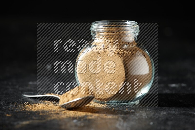 A glass spice jar and metal spoon full of allspice powder on a textured black flooring