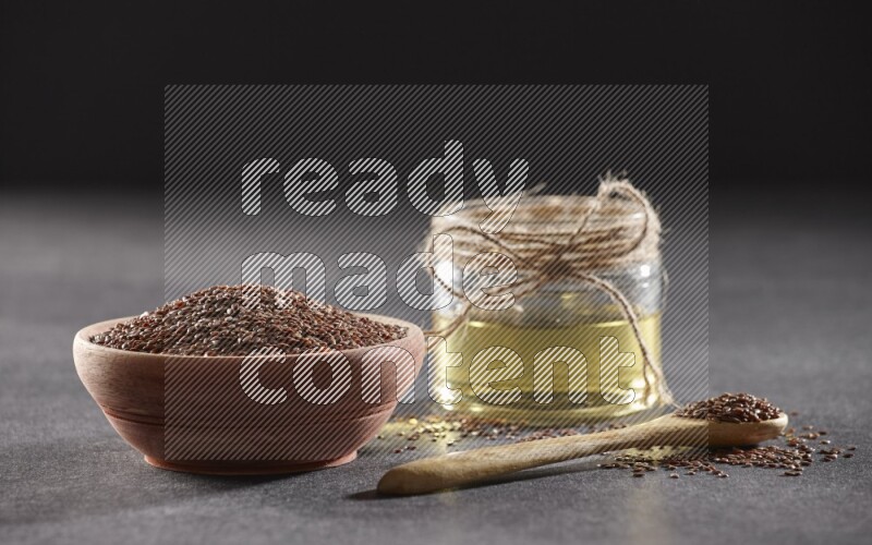 A wooden bowl and wooden spoon full of flaxseeds with a glass jar of flaxseeds oil on a black flooring