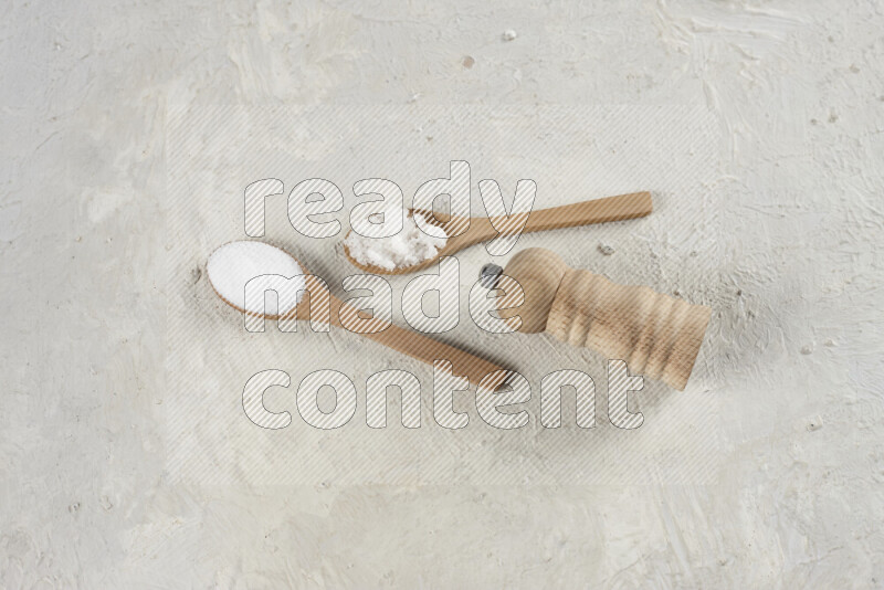2 wooden spoons filled with fine and coarse salt with wooden grinder beside them on white background