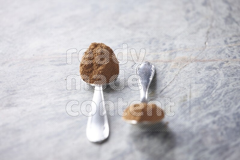 Two metal spoons full of cinnamon powder on grey marble background