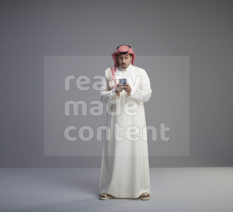 A  Saudi man standing wearing thob and red shomag texting on phone on gray background