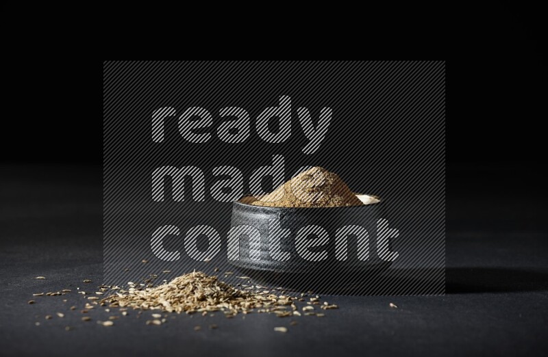 A black pottery bowl full of cumin powder and spreaded cumin seeds on a black flooring