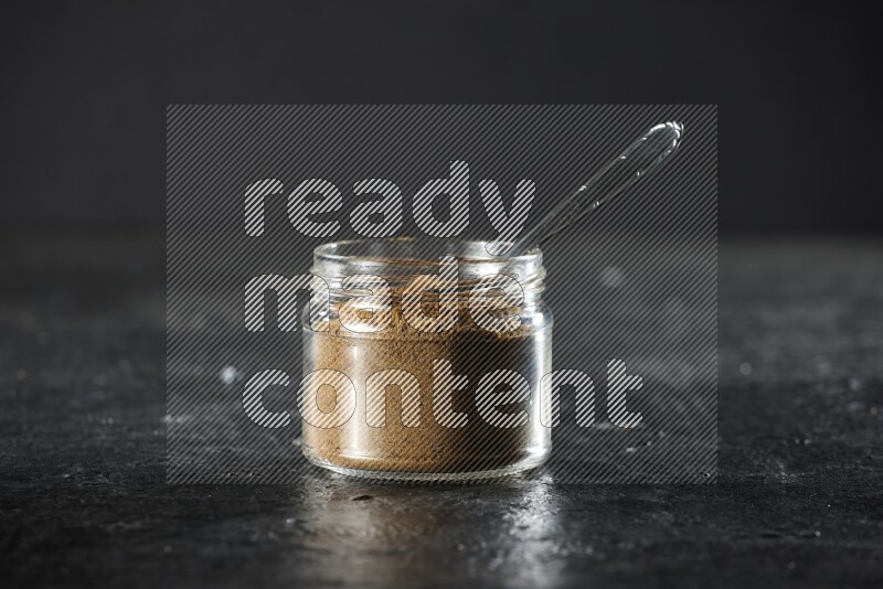 A glass jar and a metal spoon full of cumin powder on a textured black flooring
