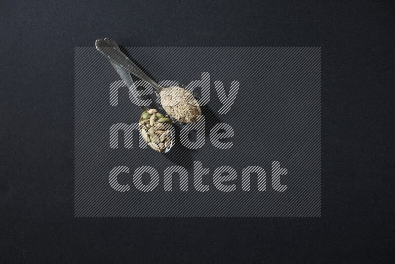 2 Metal spoons full of cardamom powder and cardamom seeds on black flooring