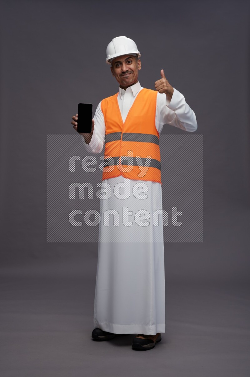 Saudi man wearing thob with engineer vest standing showing phone to camera on gray background