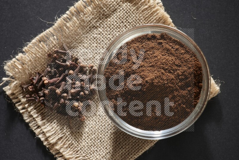 A glass bowl full of cloves powder with cloves grains on a burlap piece on a black flooring