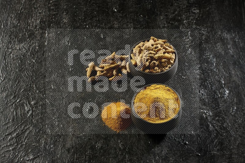 2 pottery black bowls, one full of turmeric powder and the other full of dried turmeric whole fingers on a textured black flooring