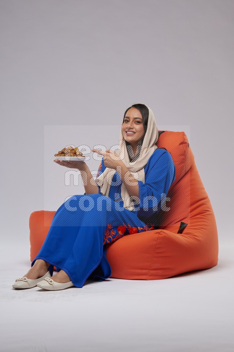 A Woman sitting on an orange beanbag wearing Jalabeya holding a plate of dates