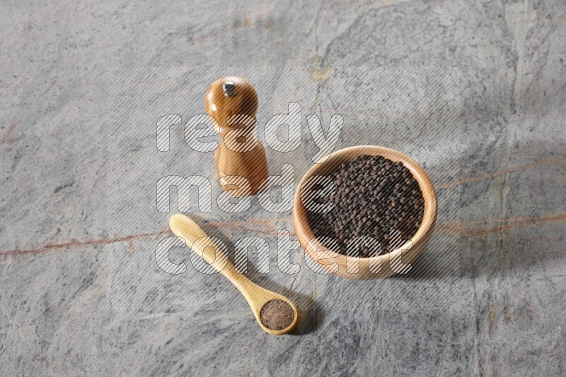 A wooden bowl full of black pepper and a wooden spoon full of black pepper powder and a wooden grinder on a marble flooring