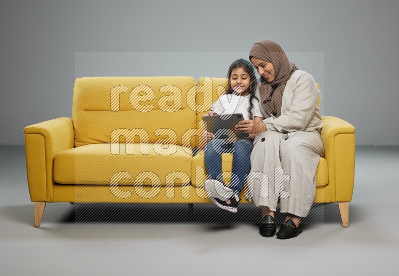 A girl with her mother on yellow sofa and watching on iPad on gray background