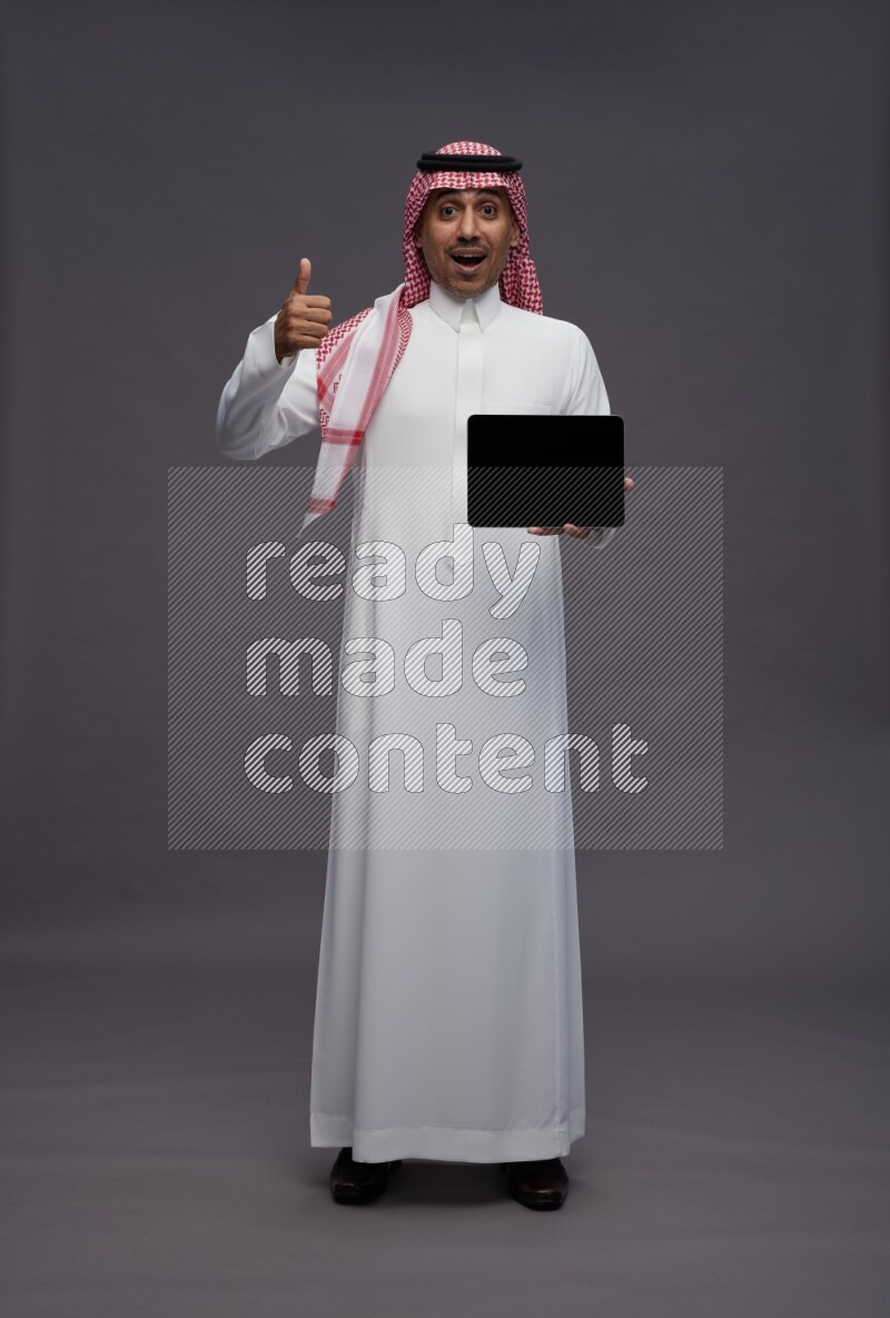 Saudi man wearing thob and shomag standing showing tablet to camera on gray background