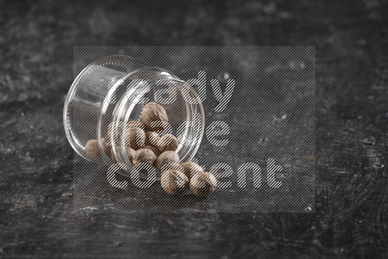 A glass jar full of nutmeg flipped and the seeds spread out on a textured black flooring