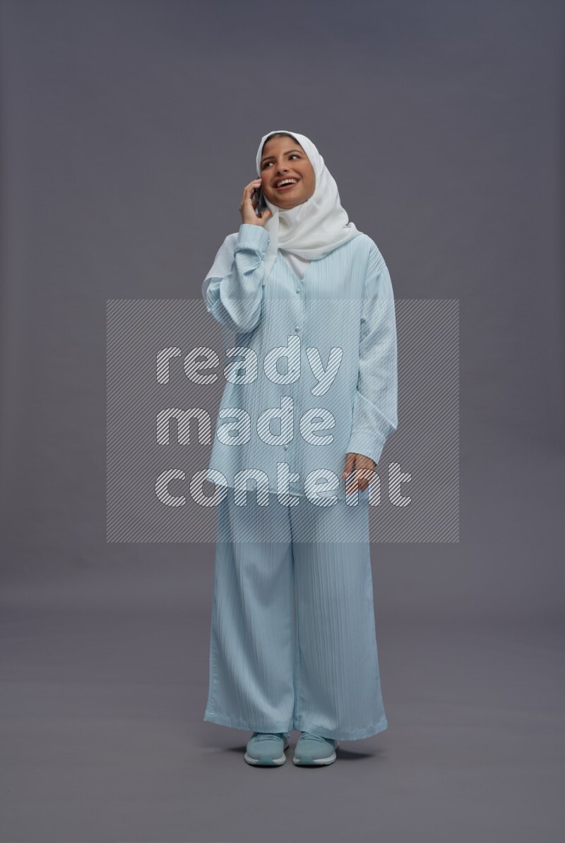 Saudi woman wearing hijab clothes standing talking on phone on gray background