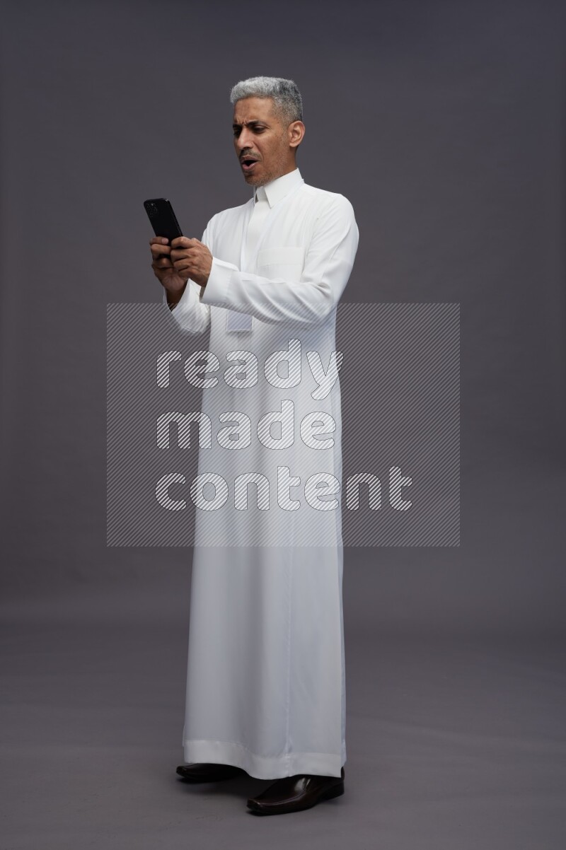 Saudi man wearing thob with neck strap employee badge standing texting on phone on gray background