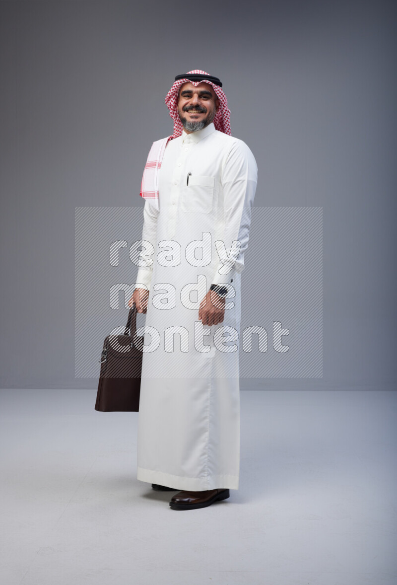 Saudi man Wearing Thob and red Shomag standing holding bag on Gray background