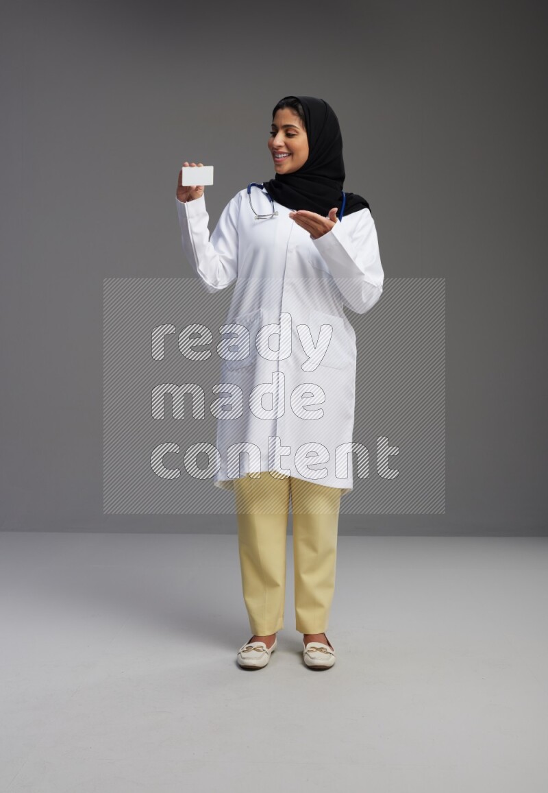 Saudi woman wearing lab coat with stethoscope standing holding ATM card on Gray background