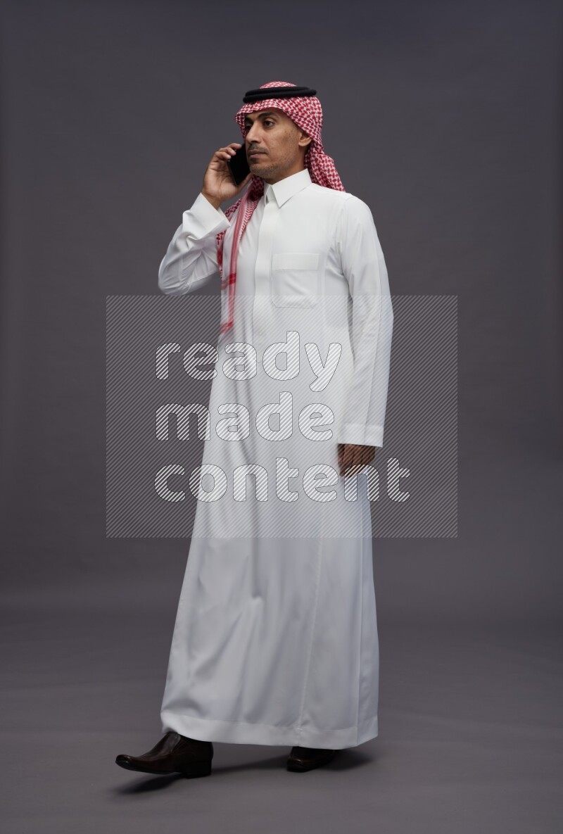 Saudi man wearing thob and shomag standing talking on phone on gray background