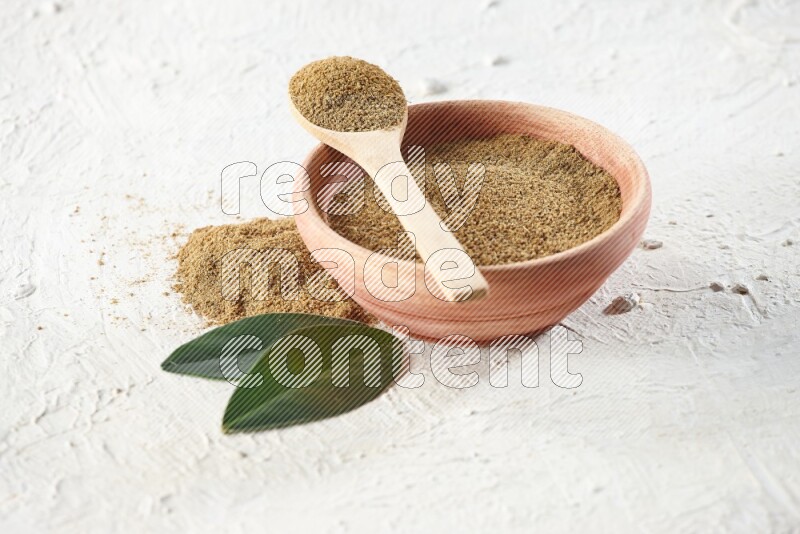 A wooden bowl and wooden spoon full of cumin powder on textured white flooring