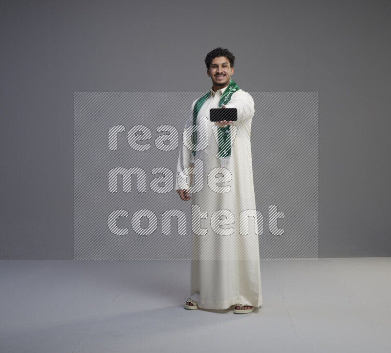 A Saudi man standing wearing thob and Saudi flag scarf showing phone to camera on gray background