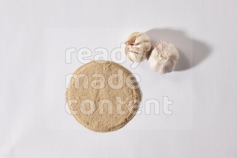 Garlic powder in a circle shape and beside it 2 garlic bulbs on a white flooring