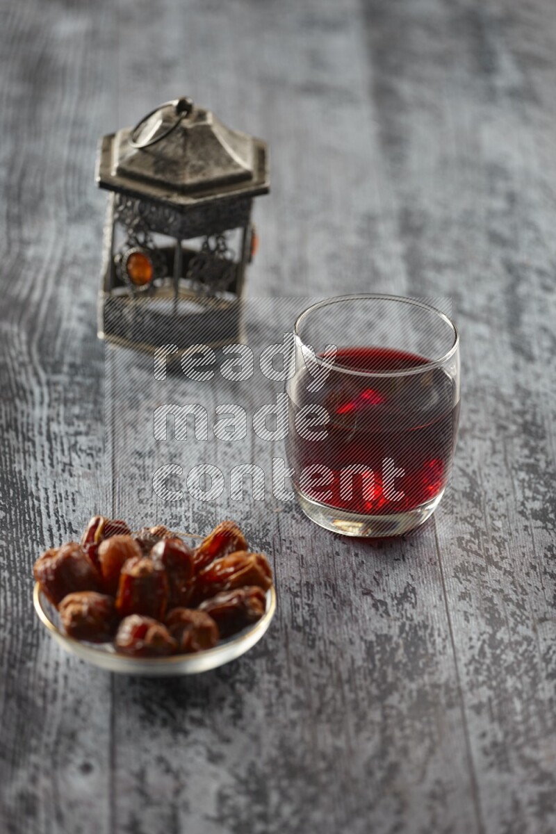 A silver lantern with different drinks, dates, nuts, prayer beads and quran on grey wooden background