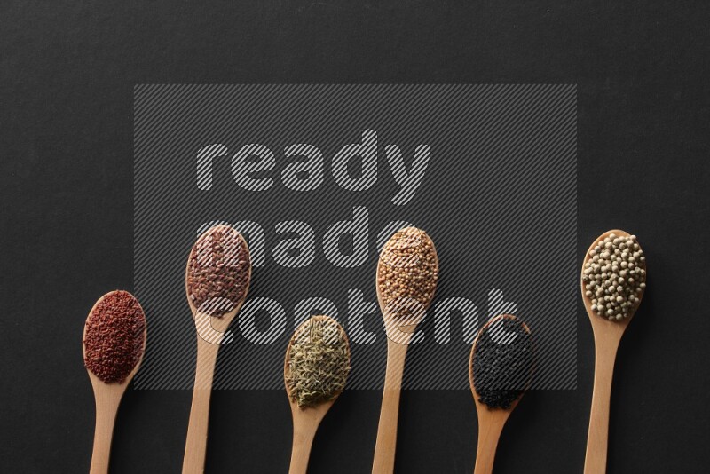 wooden spoons filled with white peppers, mustard seeds, black seeds, garden cress, cumin and flax on black flooring
