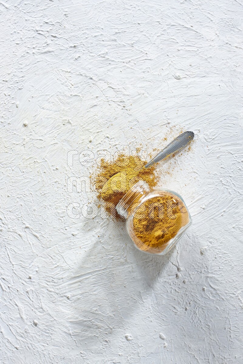 A flipped glass spice jar and a metal spoon full of turmeric powder and powder spilled out of it on textured white flooring