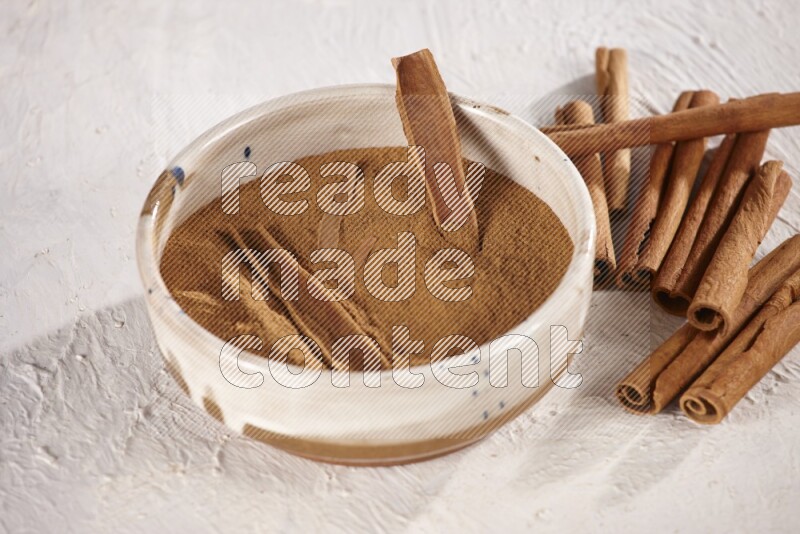 Ceramic bowl full of cinnamon powder with cinnamon sticks on the side on white background