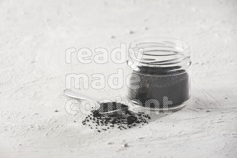 A glass jar and a metal spoon full of black seeds on a textured white flooring