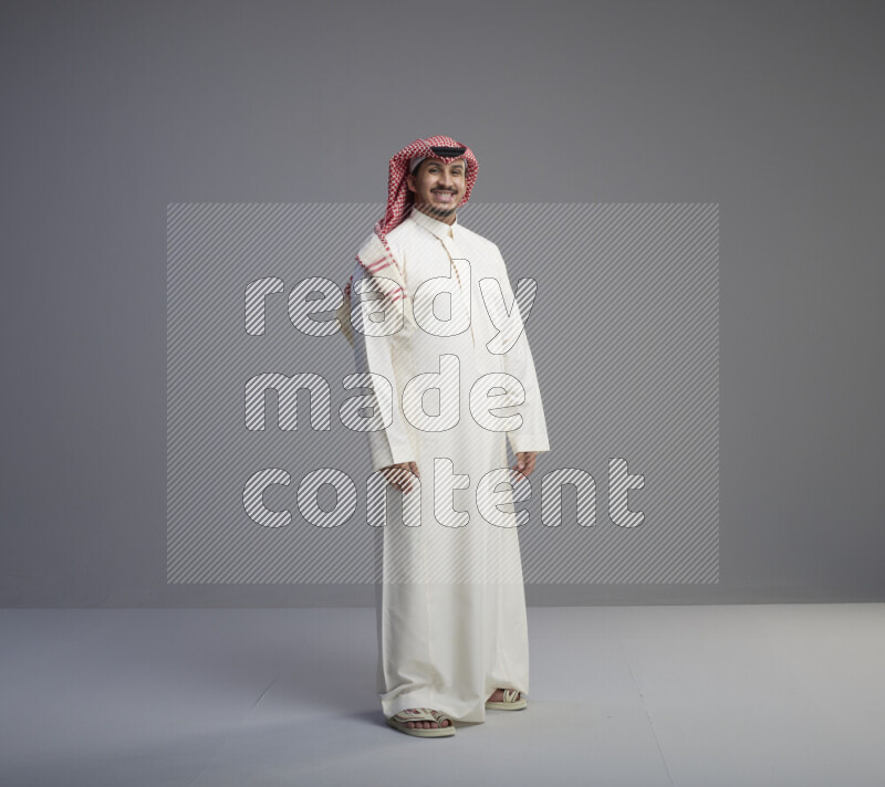 A Saudi man standing wearing thob and red shomag interacting with the camera on gray background