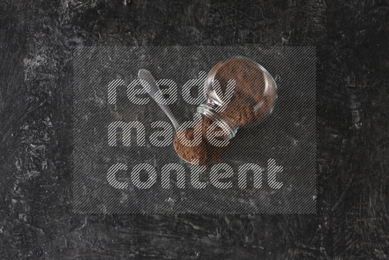 وعاء زجاجي للبهارات مقلوب وملعقة معدنية ممتلئان ببودرة القرنفل على أرضية سوداء