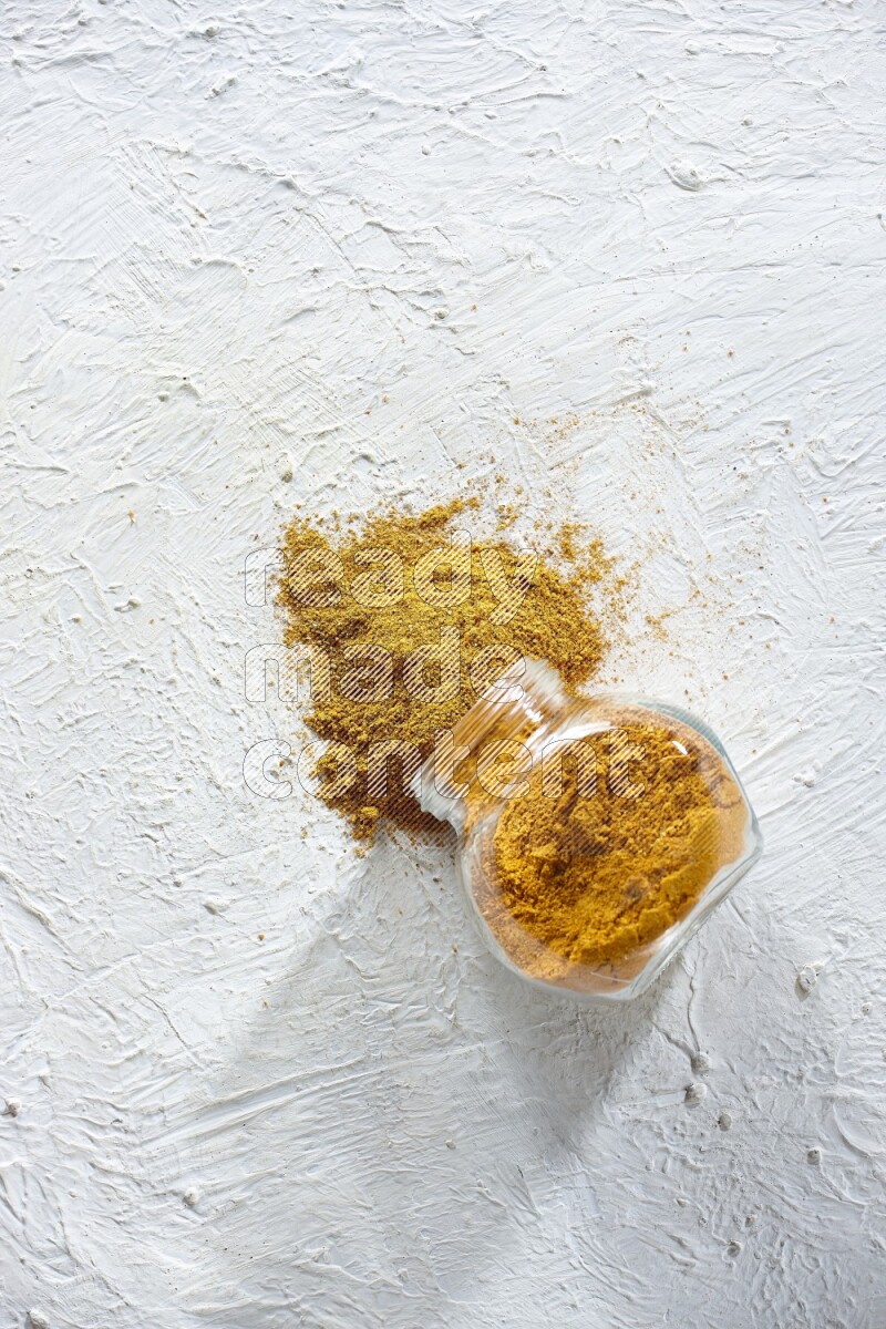 A flipped glass spice jar full of turmeric powder and powder fell out it on textured white flooring